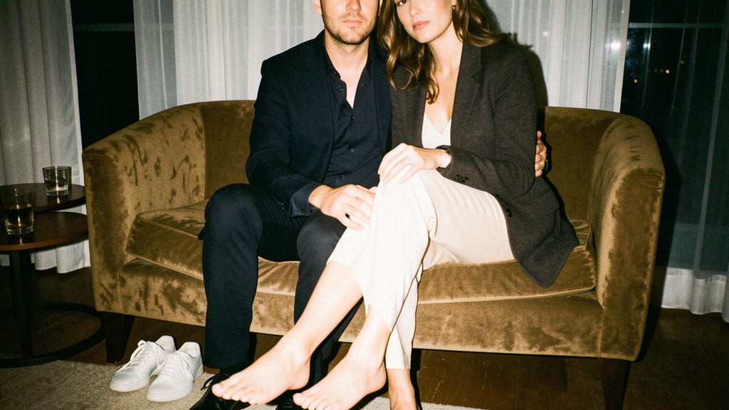 Couple seated close together on a low couch in a quiet hotel suite after hours, his jacket unbuttoned and her shoes off, faces cropped out