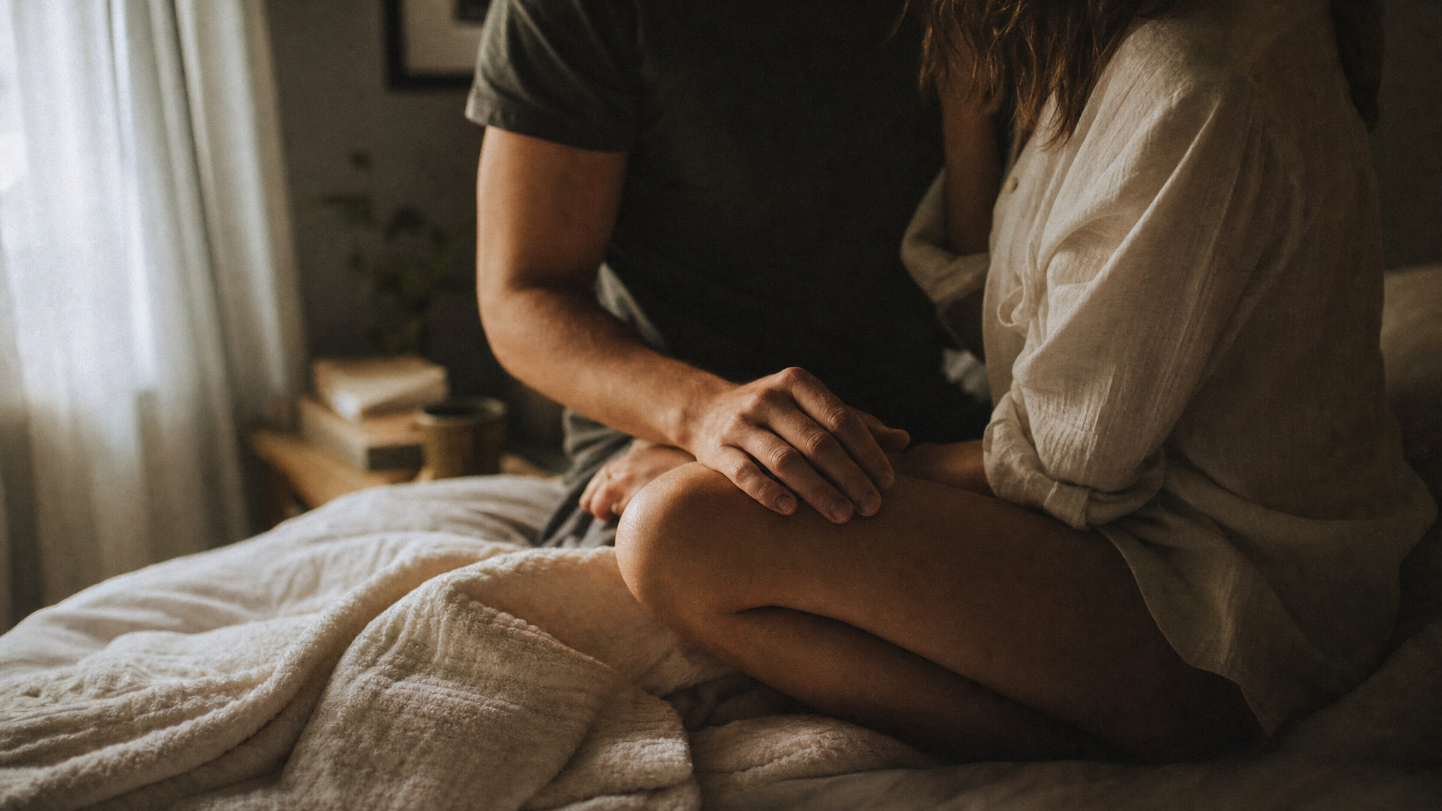 A couple sitting close on a bed in morning light, one partner's hand resting on the other's knee, quiet intimacy after a night of play