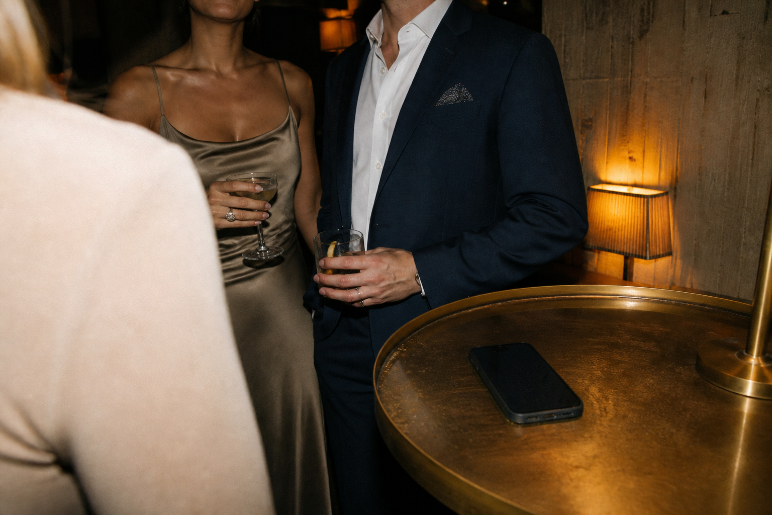 A couple in a dim private lounge at night, holding drinks and turned toward conversation off-frame, a phone face-down on the table in the foreground