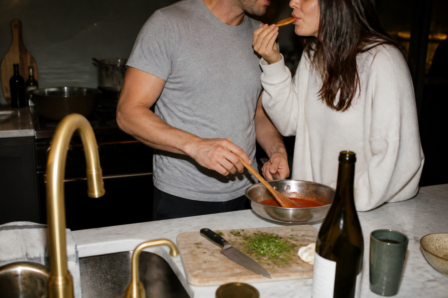 A couple standing close in a dim kitchen at night, holding wine glasses and turned toward each other in quiet conversation, warm flash-lit atmosphere